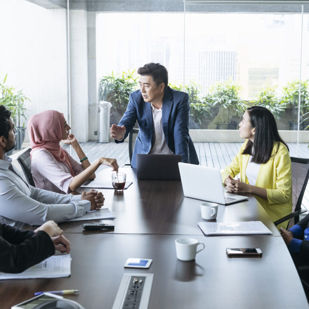 Mature businessman gesturing and looking at co-workers, multi racial group of business people sitting at table and listening